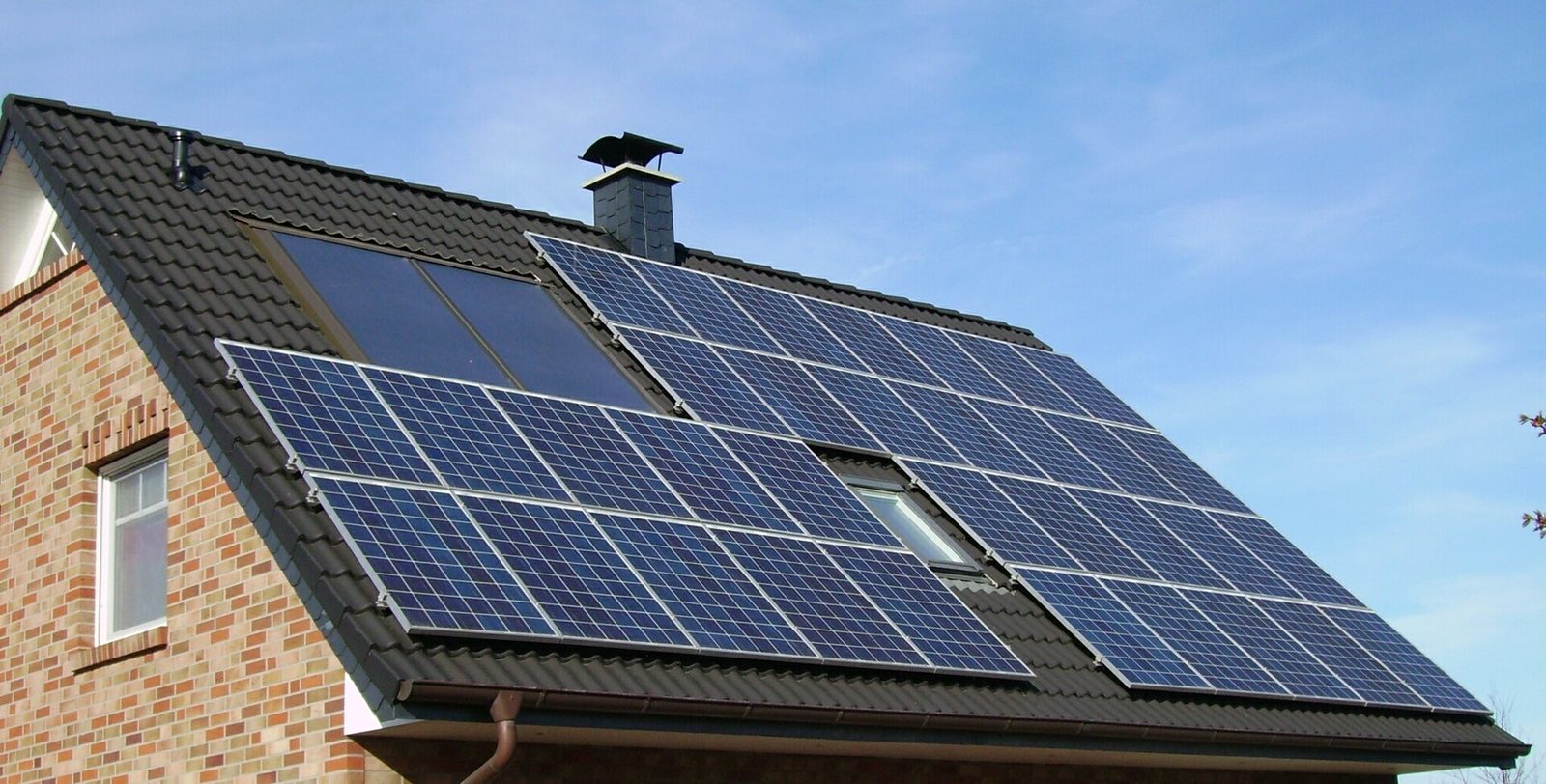 Donker pannendak van een bakstenen woning met rijen blauwe zonnepanelen en twee donkere collectoren onder blauwe lucht